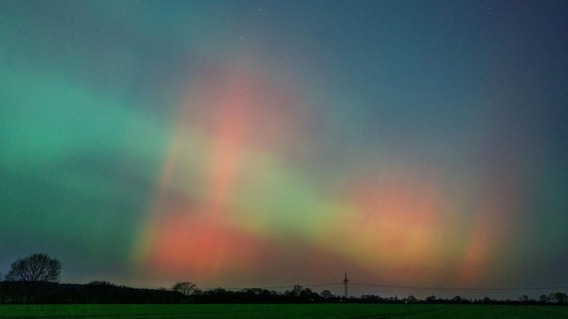 Lueur rouge et verte basse sur l'horizon au-dessus d'une plaine agricole, avec des arbres nus et un pylône électrique en silhouette