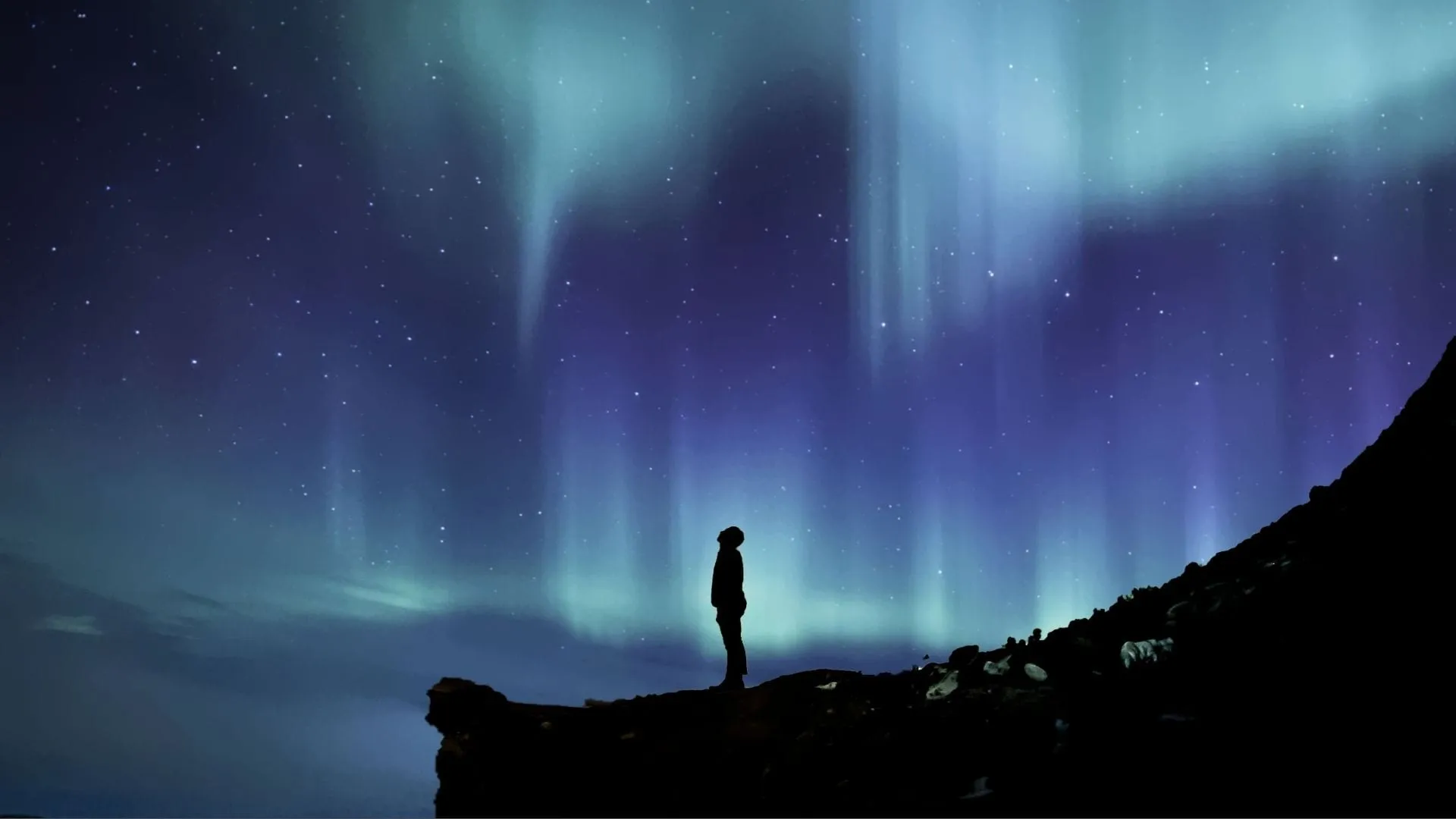 Silhouette d’une personne debout au bord d’une falaise, face à de longs rideaux d’aurore bleus et violets tombant du ciel.